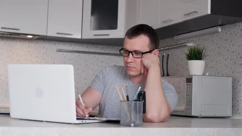 Young Man Works From Home Sitting at the Table Using Laptop Makes a Report