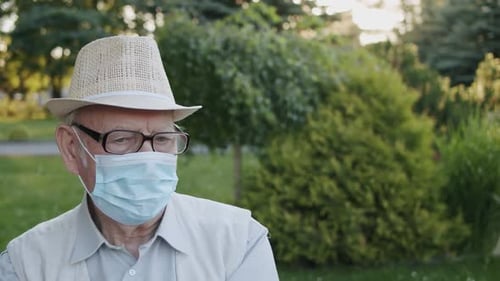 Portrait of Senior Man in Medical Mask Looking Sadly at Camera in Summer Park