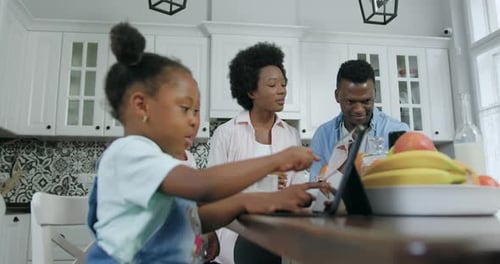 Niñas africanas sentadas frente a una tableta en la cocina mientras sus padres usan un teléfono inteligente