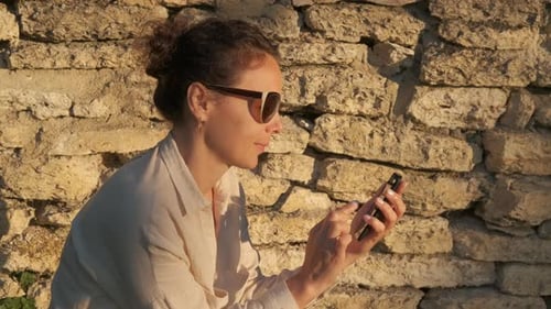 Woman Using Mobile Phone in Sunlight by Brick Wall