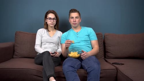 A Man and a Woman Are Watching TV. A Young Couple Are Sitting on the Couch and Eating Chips. Glass