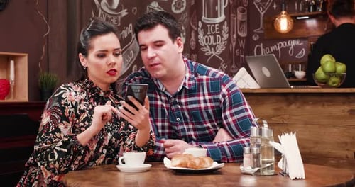 Couple on a Date in Stylish Coffee Shop Pub Restaurant Drinking Coffee and Looking on Smartphone