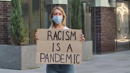 Young Woman in Medical Mask Stands with a Cardboard Poster RACISM IS A PANDEMIC in a Public Place