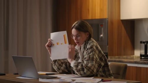 Home Office A Young Woman with Glasses Shows a Graph to a Laptop Camera While Sitting at Home in the