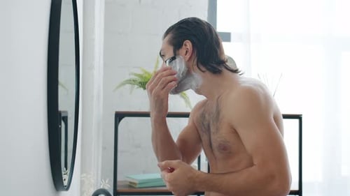 Slow Ralenti d'un gars se rasant le visage avec un rasoir moderne regardant un miroir dans la salle de bain