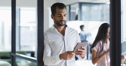 Mixed race businessman standing in office doorway, using tablet, smiling to camera
