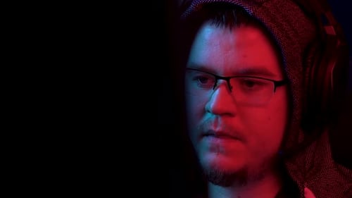 A Young Man Sits at a Computer with Headphones. A Man in a Hood Looks at the Monitor and