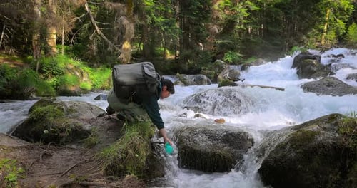Female Traveler with a Backpack Drinking Water in Nature in the Forest Near a Mountain River