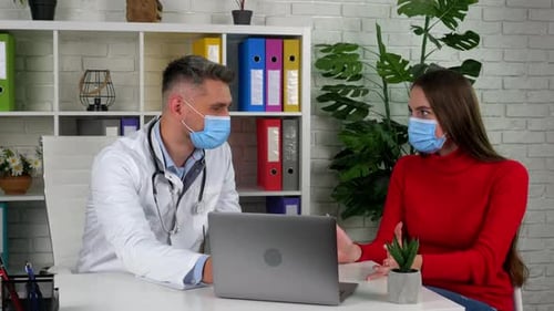 Doctor and patient wear protective masks on faces in hospital office talk listen