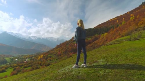 woman in denim jacket is standing on top of mountain, raising her hands in air,
