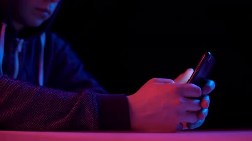 Young Man in a Hood with a Smartphone. Hacker Makes a Hack Through the Phone. Blue and Red Light