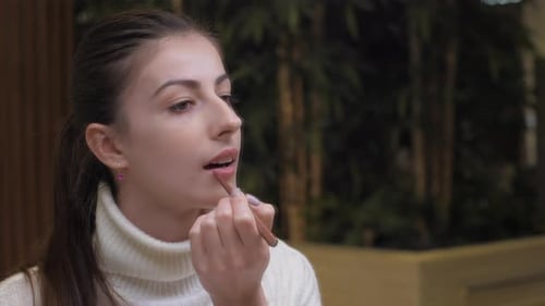 Une jeune femme en pull blanc applique un crayon à lèvres rouge mat