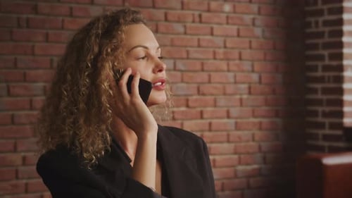 Young Adult Curly Blond 30s Businesswoman is Talking on a Phone in a Loft Apartment