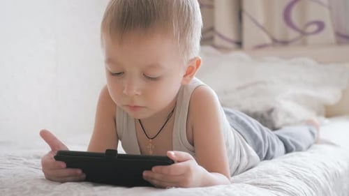 Little Boy Sitting on a Bed and Plays on the Tablet in the Room