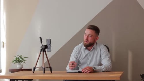 Man with a Beard in a Blue Shirt Sits at His Desk in Room Blogs on Phone Online