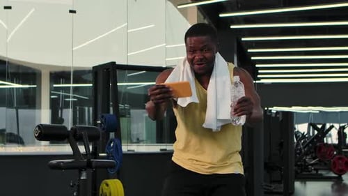 Black Man Looks at Phone in Gym While Drinks Water While Relaxing Then Celebrates Victory and
