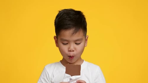 Little Japanese Boy Eating Chocolate Bar Over Yellow Studio Background