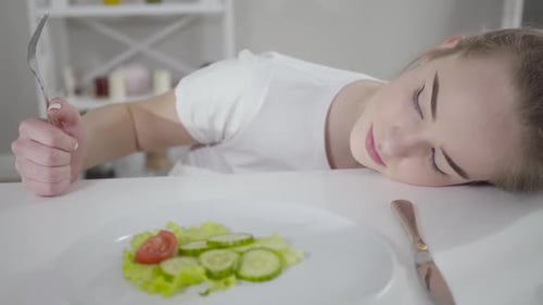 Verärgertes kaukasisches Mädchen mit Gabel, das auf dem Tisch liegt und Salat aus geschnittenen Tomaten und Gurken betrachtet