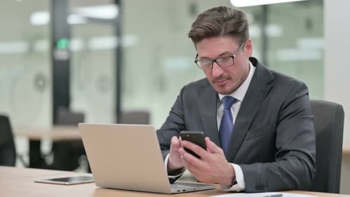 Middle Aged Businessman Working on Laptop and Smartphone in Office