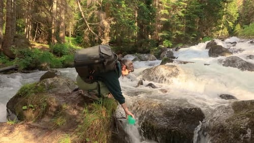 Female Traveler with a Backpack Drinking Water in Nature in the Forest Near a Mountain River