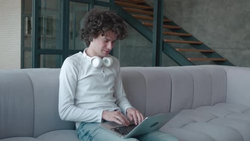 Young Guy From the Tunnels in the Ears in Leaning on Comfortable Sofa Working Remotely on Computer
