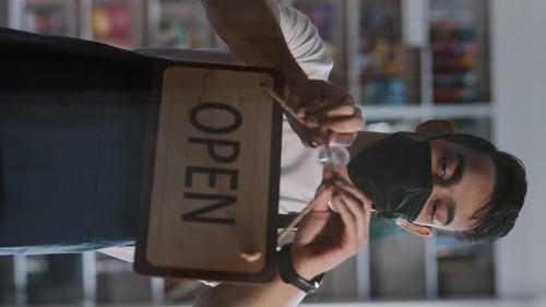 Young Successful Male Owner in Medical Mask Stands Behind Glass Door Establishment and Turns Sign