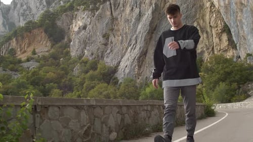 A Young Stylish Man Walks Along a Mountain Road and Throws Stones From a Cliff