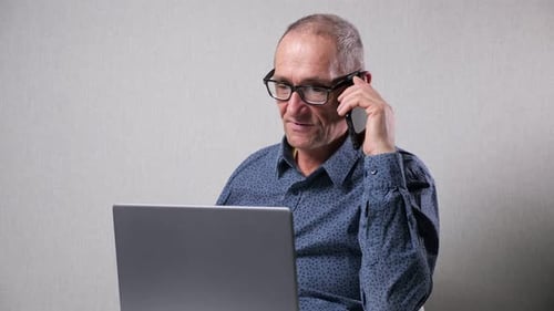 Älterer Mann spricht am Handy und arbeitet am Laptop am Tisch