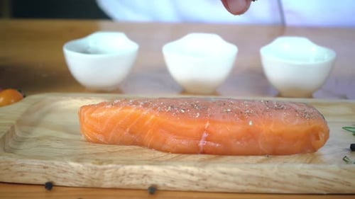 Chef cook season fresh chunk of deli piece of salmon fish with sea salt and grounded spicy peppers.