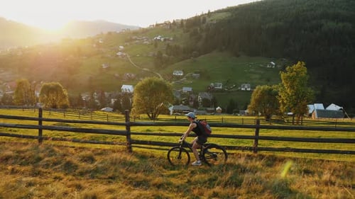 Young Woman Rides a Bicycle Along a Wooden Fence High in the Mountains with a View of the Village