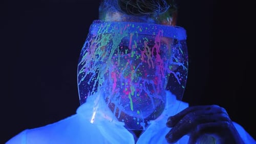 Man Removing Paint Splattered Face Shield Under Blue Light