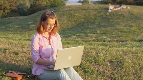 A young woman works on a laptop remotely while sitting in a park in nature in the summer.