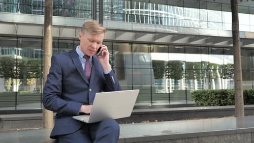 Businessman Talking on Phone while Working on Laptop Outside Office