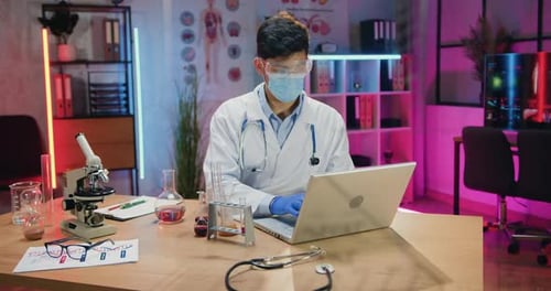 Homme scientifique portant un masque de protection travaillant sur un ordinateur portable pendant des heures supplémentaires dans un laboratoire du soir