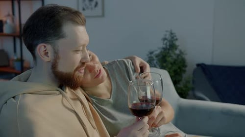 Junger Mann vor dem Fernseher mit Freundin trinkt Rotwein und spricht auf der Couch im dunklen Haus