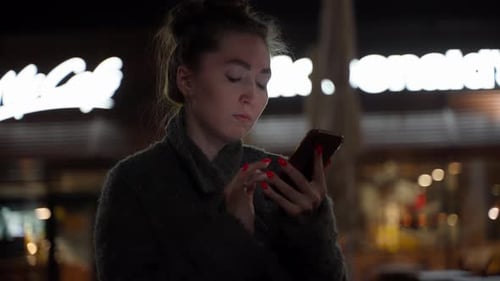 Attractive Young Hipster Woman in Coat Using Mobile Phone Standing on Street of Night Town Reacting