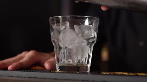Closeup Cropped Shot of Unrecognizable Barman Pouring Mixed Cocktail Into Glass with Ice Cube on Bar