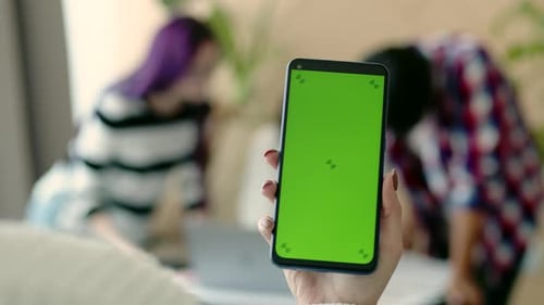 Woman's hand holds phone with green screen against the background of working team of people. Mockup,