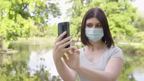 A Young Caucasian Woman in a Face Mask Takes Selfies with a Smartphone in a Park