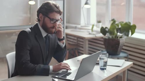 Young Businessman Having Phone Talk and Using Laptop at Office Desk
