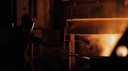 Steel Worker in Protective Clothing raking Furnace in an Industrial Foundry.