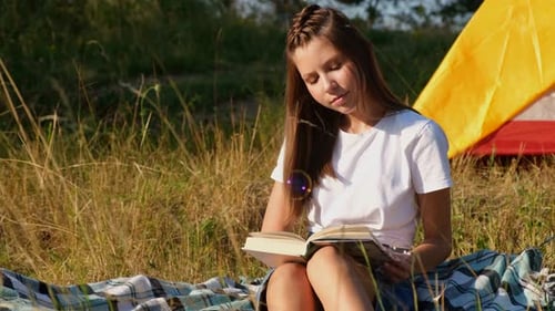 A cute little girl is reading a book at a summer camp. Tent camp.
