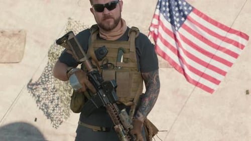 Man Wearing Vest Holding Rifle in Front of Flag