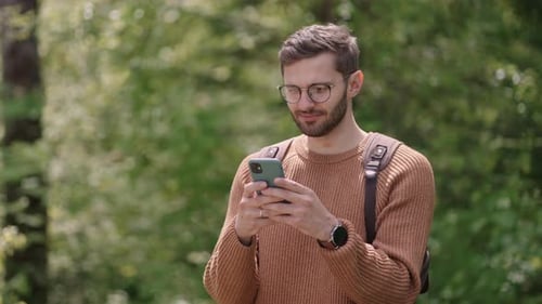 Smiling Man with Glasses with a Beard Walks Through the Woods with a Backpack and Prints a Message
