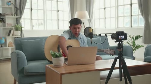 Chico asiático vlogger con guitarra buscando comentarios en una computadora portátil. El chico está transmitiendo