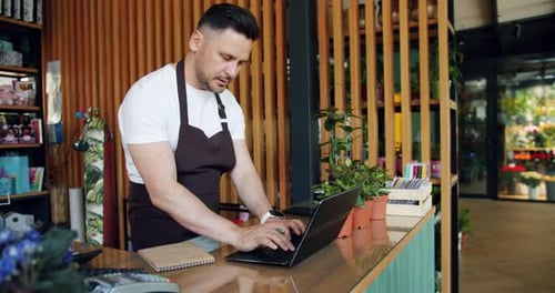 Entrepreneur Working with Laptop in Flower Shop Typing and Writing in Notebook