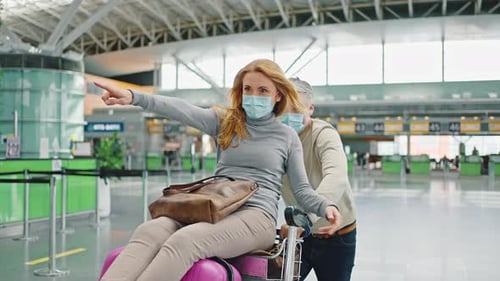 Happy Mature Couple in Protective Masks Fooling Around Waiting Departure Riding on Luggage Cart at