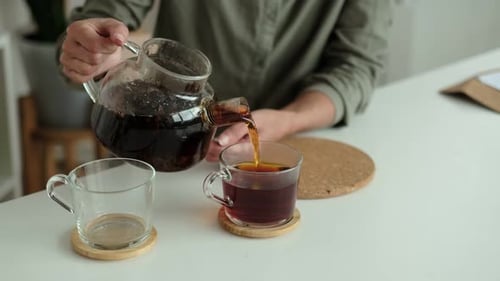 Frau in einem Leinenhemd gießt Tee in zwei durchsichtige Tassen