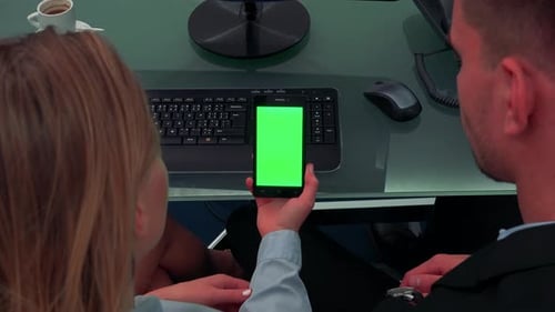 A Man and a Woman Sit at a Desk in an Office and Look at a Smartphone with a Green Screen