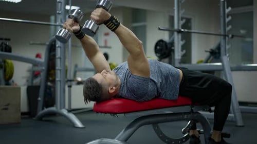 Starker männlicher Sportler, der auf Gymnastikbank liegt und Hanteln hebt